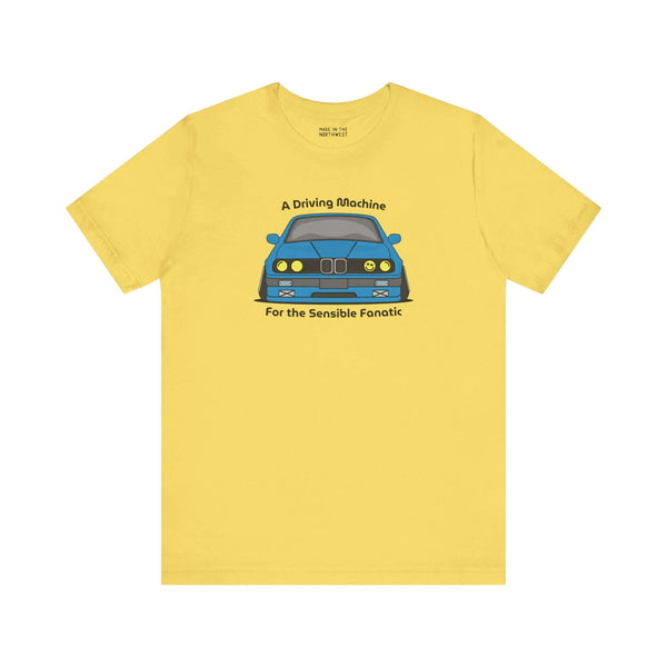 Yellow soft tee featuring the illustration of a BMW E30 with the text 'A Driving Machine for the Sensible Fanatic'.