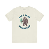 Don’t Be an Assquatch Tee featuring Bigfoot holding flowers and humorous text in a light-colored t-shirt.