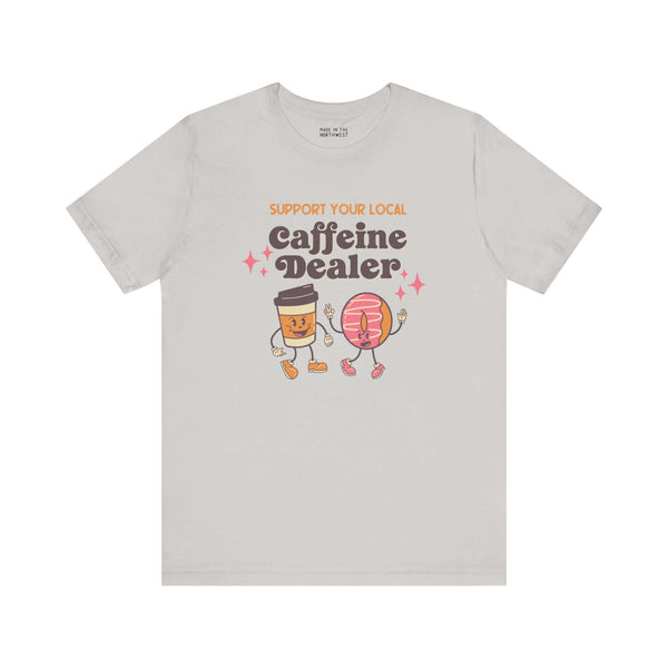 "Support Your Local Caffeine Dealer coffee tee with fun graphic design, featuring coffee cup and donut characters on a soft gray fabric."