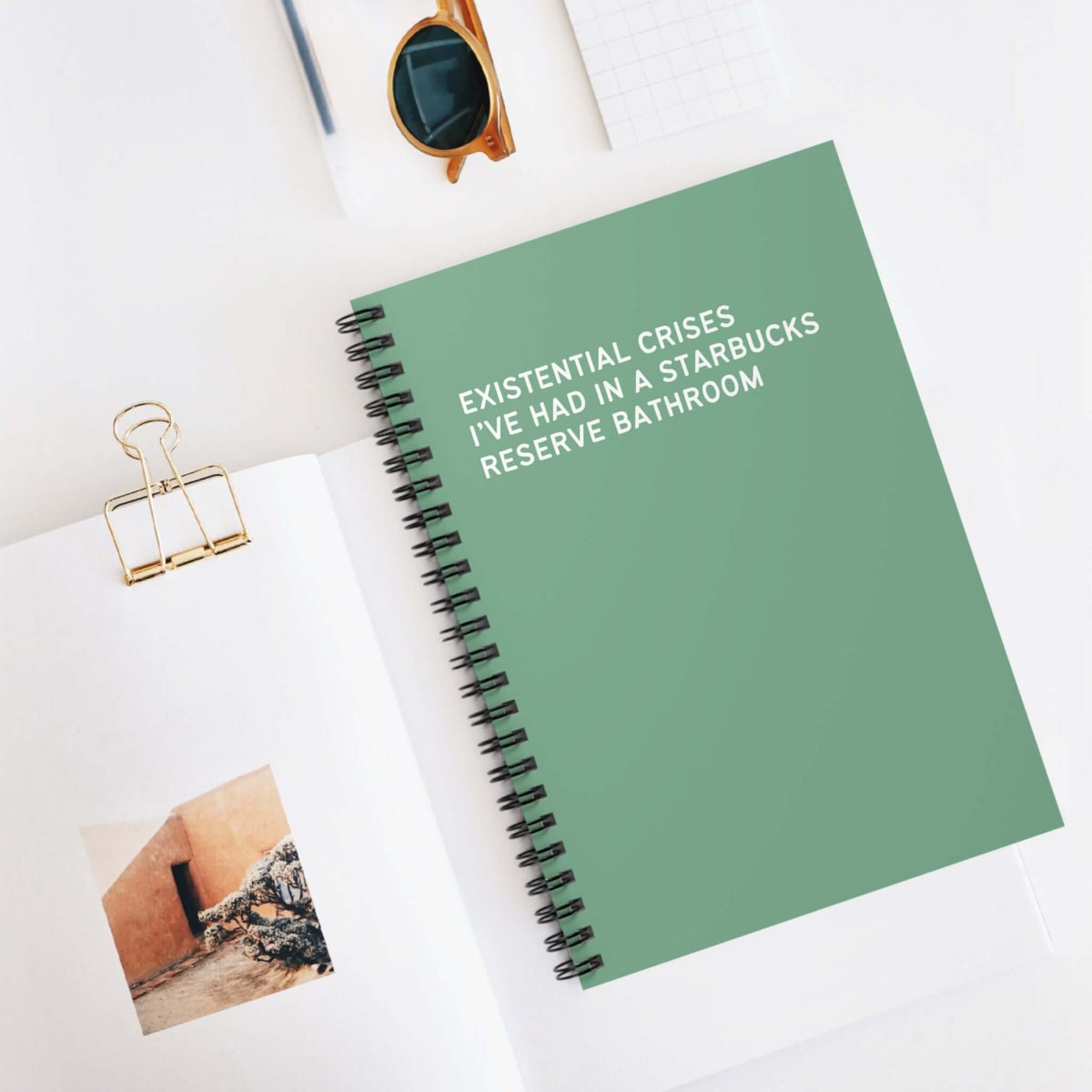 Green spiral notebook titled 'Existential Crises I've Had in a Starbucks Reserve Bathroom' on a desk.