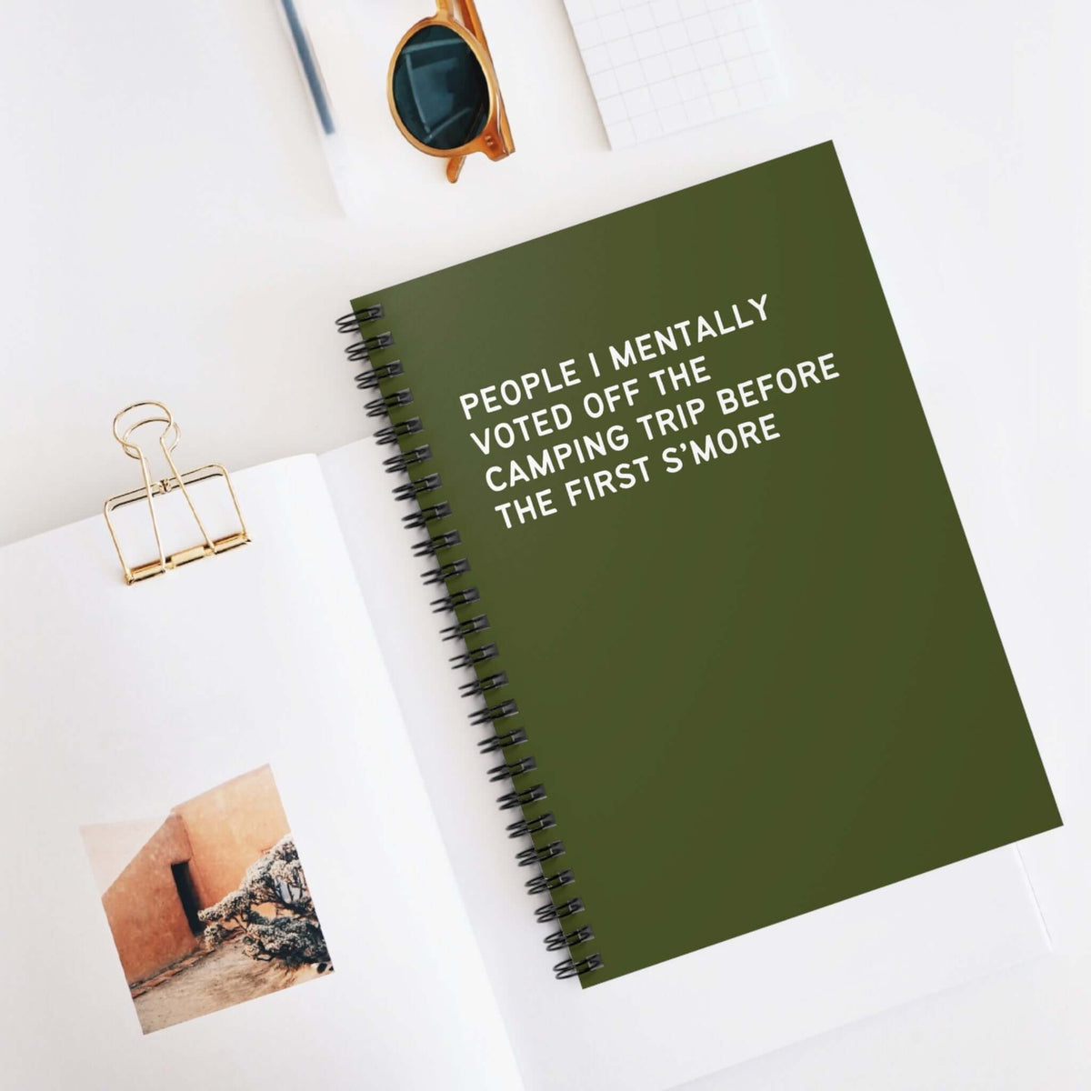 Green spiral notebook titled 'People I Mentally Voted Off the Camping Trip' on a desk with sunglasses.