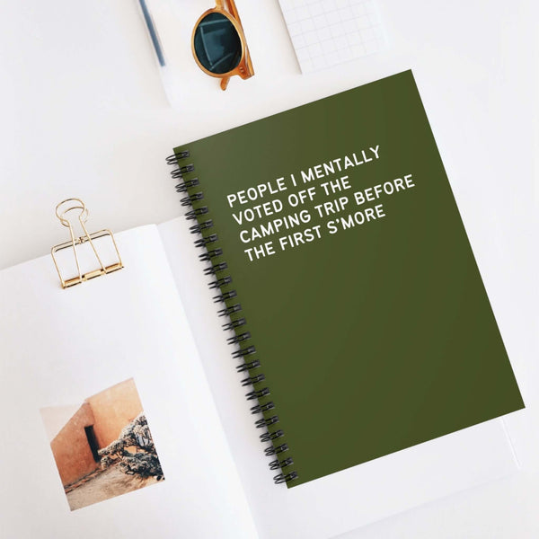 Green spiral notebook titled 'People I Mentally Voted Off the Camping Trip' on a desk with sunglasses.