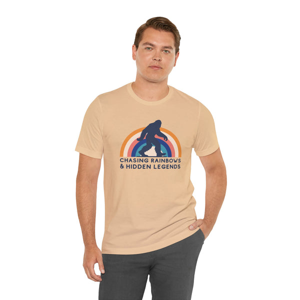 Man wearing Chasing Rainbows and Sasquatch Premium Soft Tee with colorful graphic on a beige shirt.