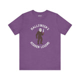 Halloween's Hidden Legend Bigfoot tee features a creature in a mask holding a knife on purple fabric.