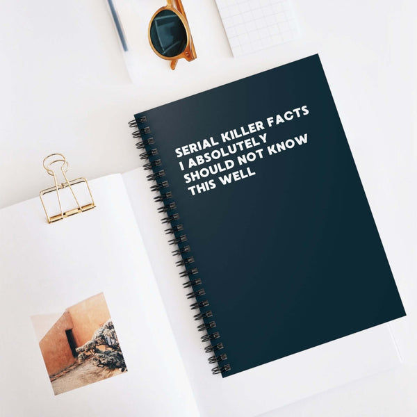 Spiral notebook titled 'Serial Killer Facts I Absolutely Should Not Know This Well' on a desk with sunglasses and a photo.
