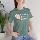 A smiling person wearing a Serving Holiday Cheer Pickleball T Shirt, surrounded by greenery.