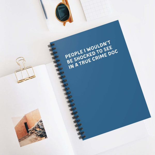 Spiral notebook titled 'People I Wouldn’t Be Shocked to See in a True Crime Doc' on a desk with other stationery.