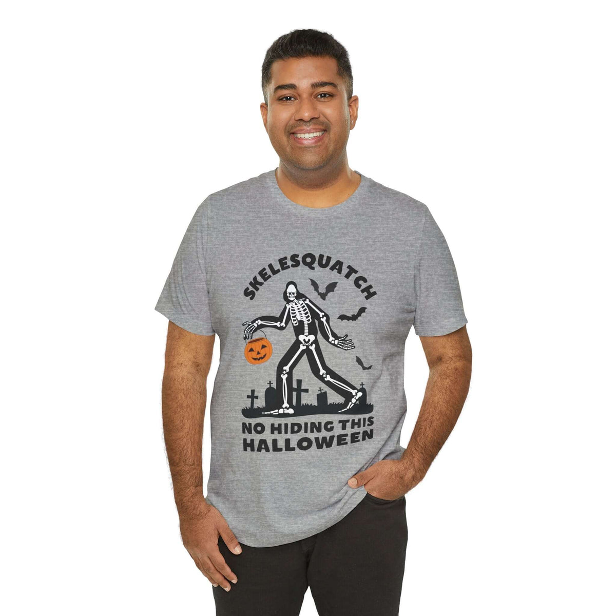 Man wearing Skelesquatch Halloween tee, featuring a skeleton Sasquatch with a pumpkin bucket in a graveyard design.