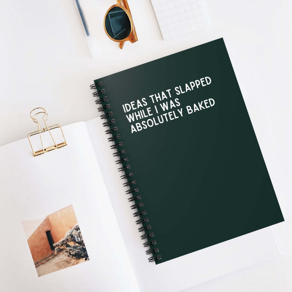 Spiral notebook titled 'Ideas That Slapped While I Was Absolutely Baked' on a stylish desk with sunglasses.