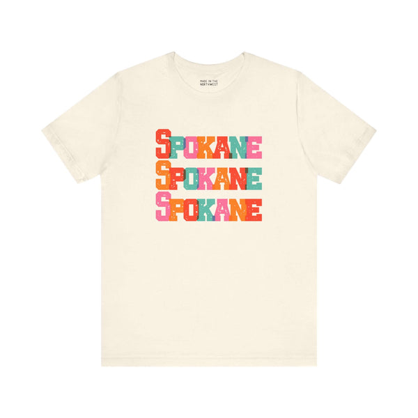 Cream tee with bold, colorful "Spokane" text design, perfect for fans of Lilac City.