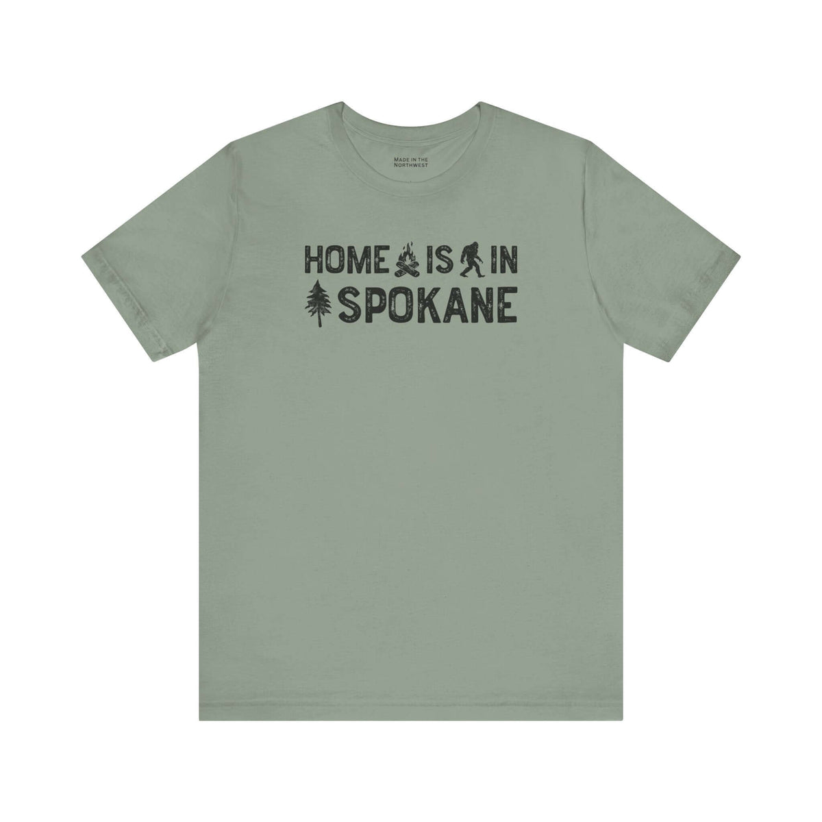 Home Is in Spokane Tee featuring text and illustrations of Bigfoot, a tree, and a campfire on a green shirt.