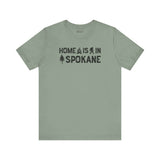 Home Is in Spokane Tee featuring text and illustrations of Bigfoot, a tree, and a campfire on a green shirt.