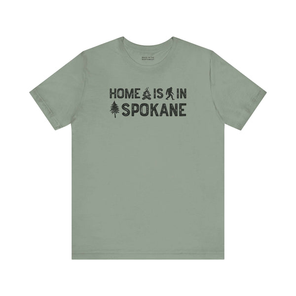Home Is in Spokane Tee featuring text and illustrations of Bigfoot, a tree, and a campfire on a green shirt.