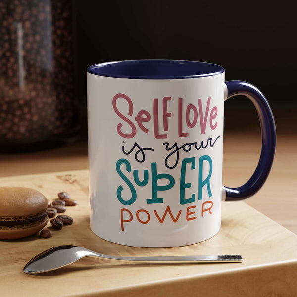 Self Love Is Your Super Power Mug with colorful text, navy interior, on a wooden table with a spoon and macaron.