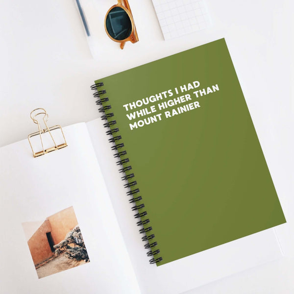 Green spiral notebook titled 'Thoughts I Had While Higher Than Mount Rainier' on a stylish desk.