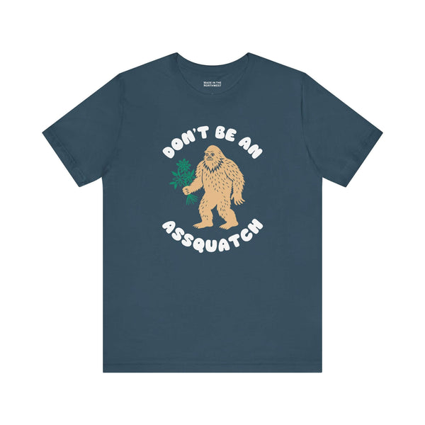 Don’t Be an Assquatch Tee featuring a friendly Bigfoot holding flowers on a navy shirt.