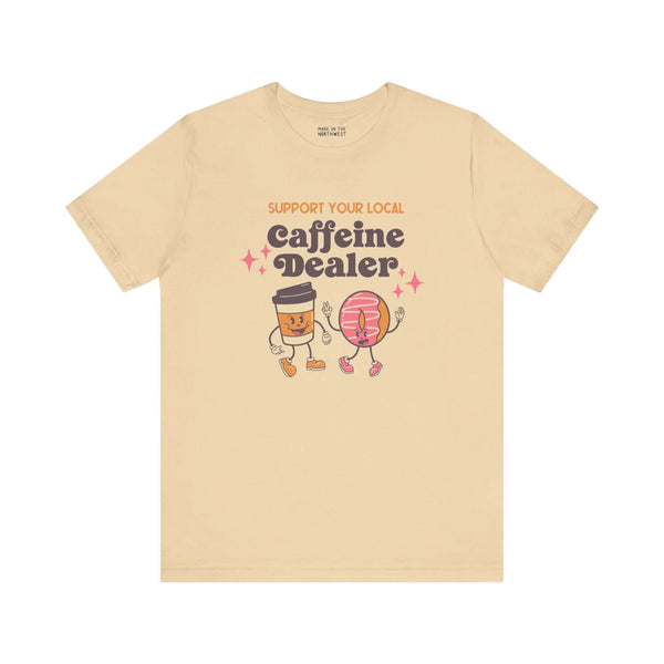 "Beige coffee-themed tee with 'Support Your Local Caffeine Dealer' design featuring playful coffee cup and donut characters."