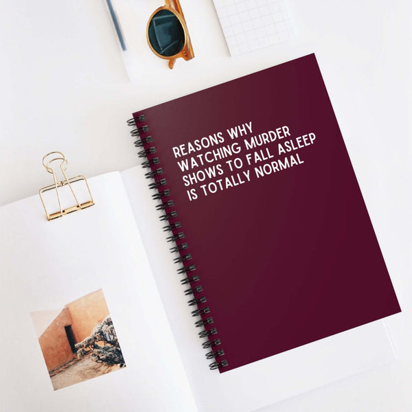 Spiral notebook titled 'Reasons Why Watching Murder Shows to Fall Asleep is Totally Normal' on a desk.