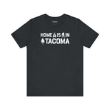 Home is in Tacoma tee featuring campfire, tree, and Bigfoot symbols on a black t-shirt.